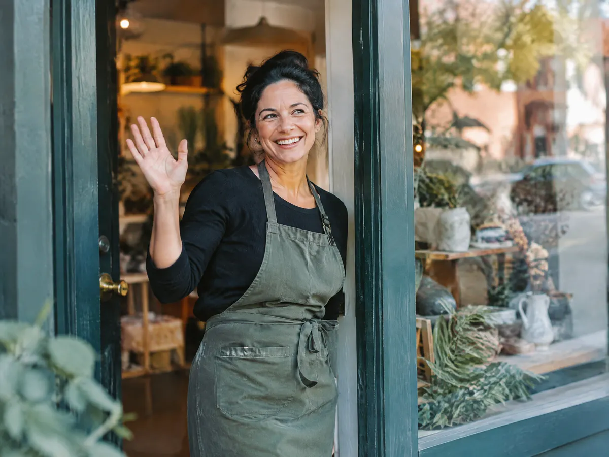 A small business owner waving from the door of her shop.