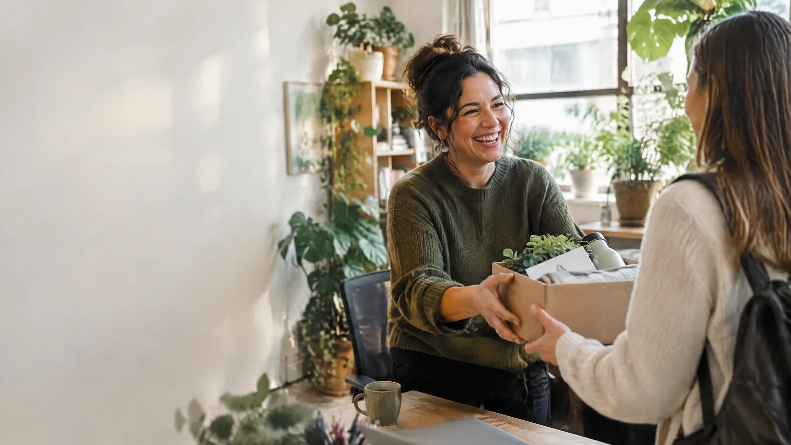An office manager in a plant-filled small office laughing with a new hire as she hands over a welcome package.