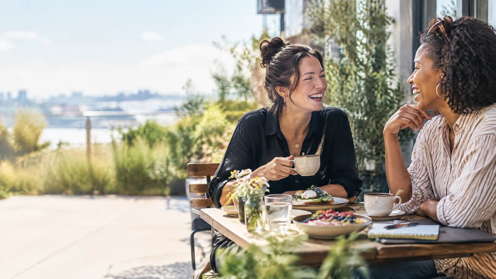 Two friends who both run small businesses chatting over brunch at a sunny patio, comparing notes, laughing.
