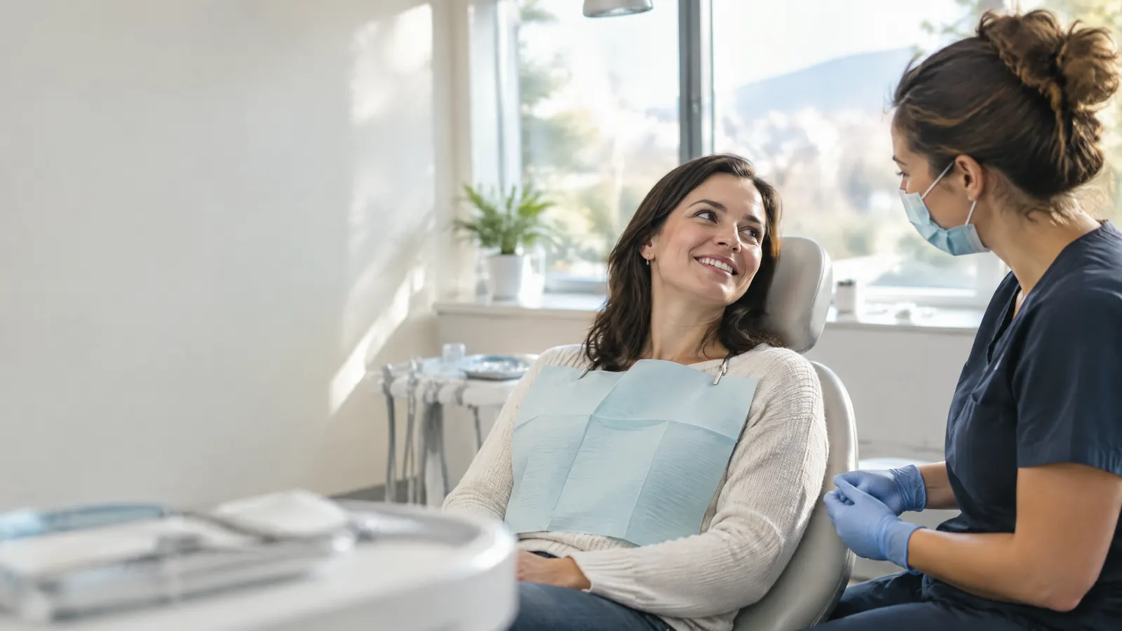 A relaxed woman in her thirties smiles while a dental hygienist in scrubs and a mask chats with her beside a bright, modern treatment chair with soft daylight from a window.