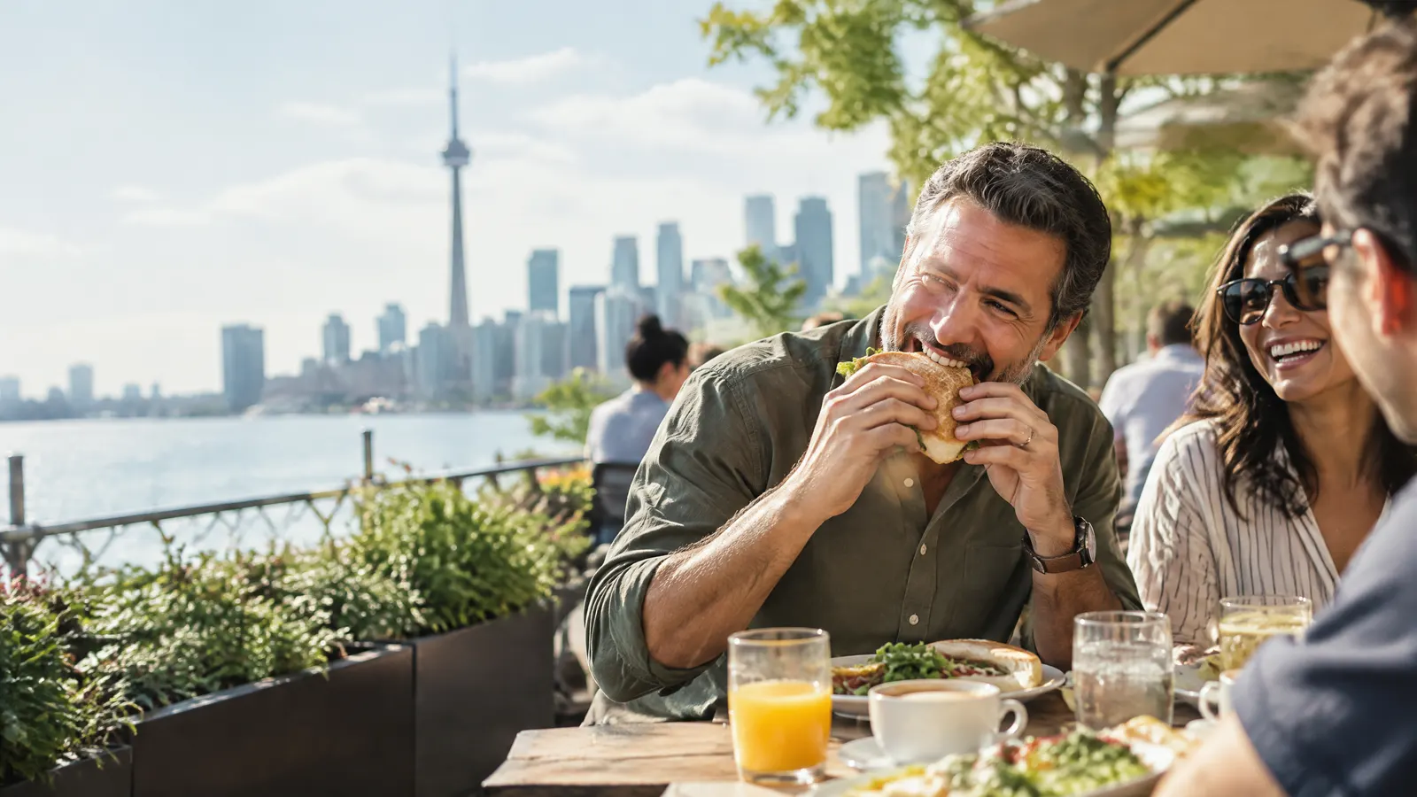 A middle-aged man laughs over brunch with friends on a sunny Toronto patio, biting confidently into a crusty sandwich.