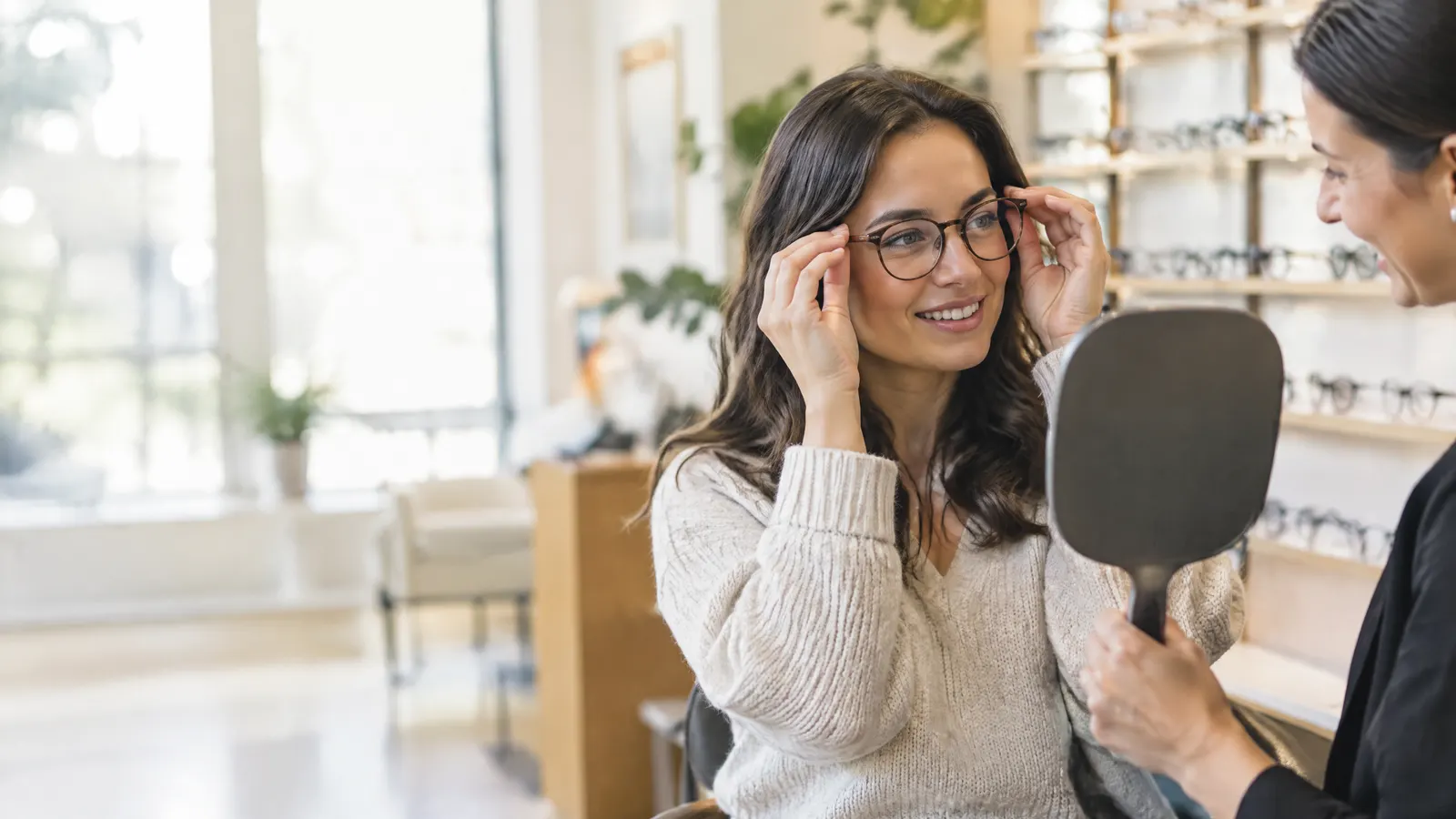 A woman in her thirties tries on a pair of round frames in a bright optical shop while an optician holds up a hand mirror and smiles.