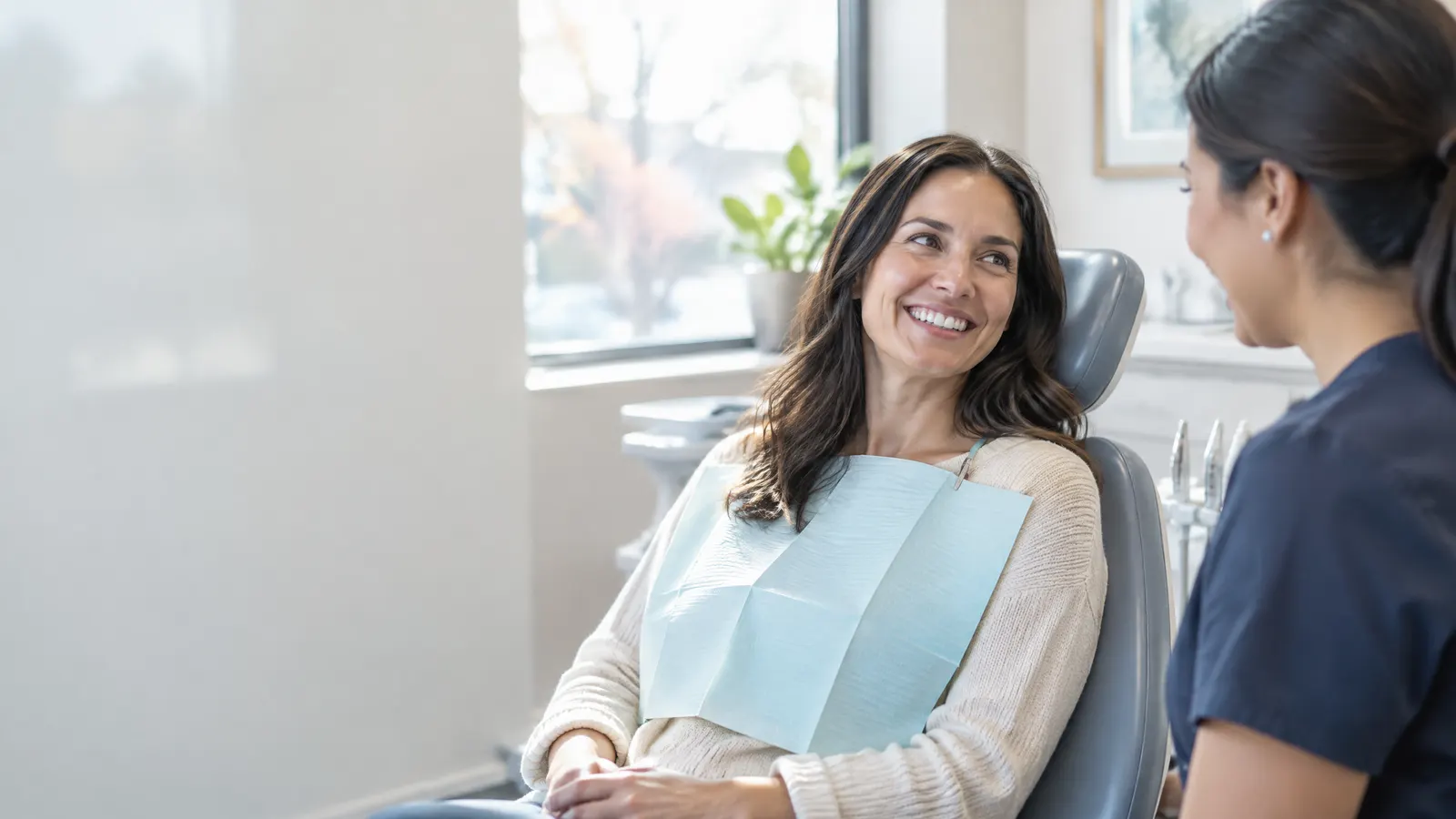 A smiling woman in her thirties sits in a bright dental office chair chatting with her hygienist after a cleaning, with soft daylight through a window.