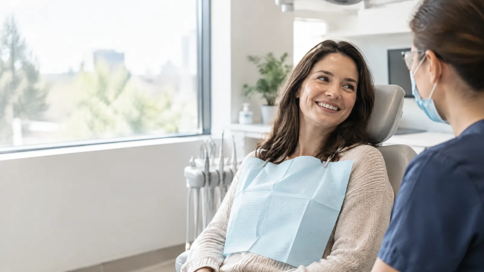 A relieved woman in her thirties smiles gently while chatting with her dentist in a bright, modern dental office after a treatment, natural daylight through a window.