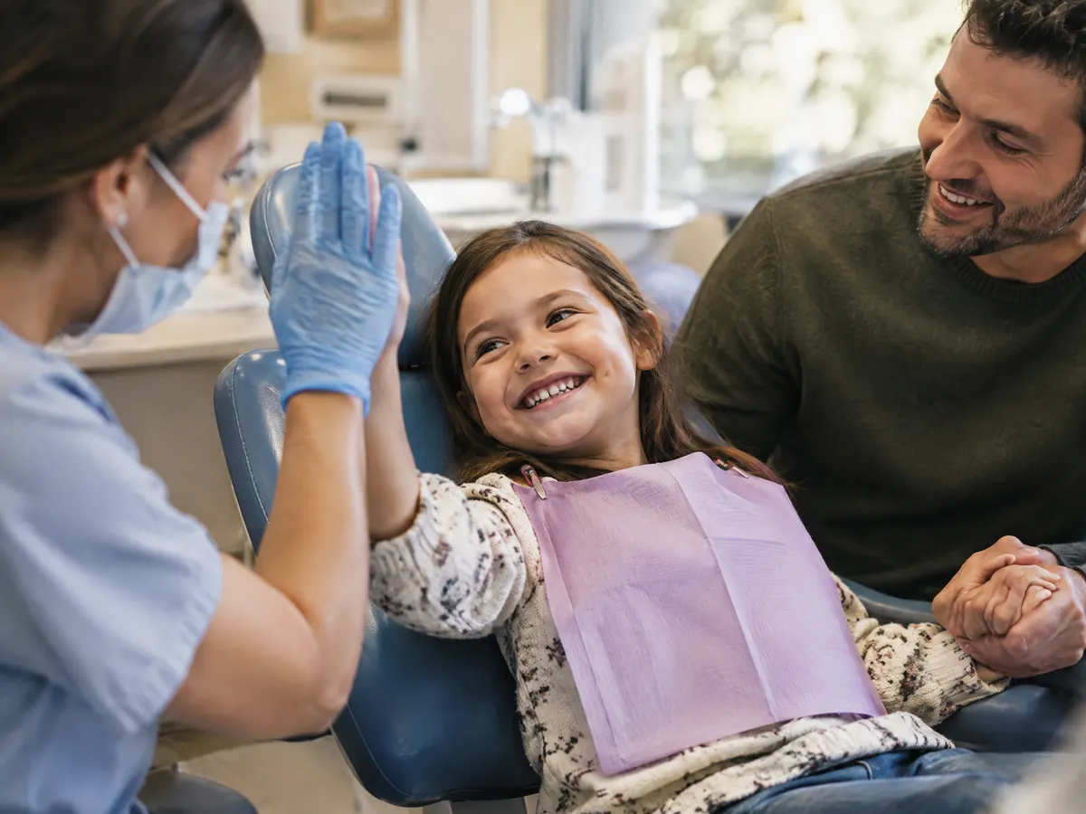 A young girl giving a big smile in a dental chair while her dad holds her hand and a dentist gives a high five.