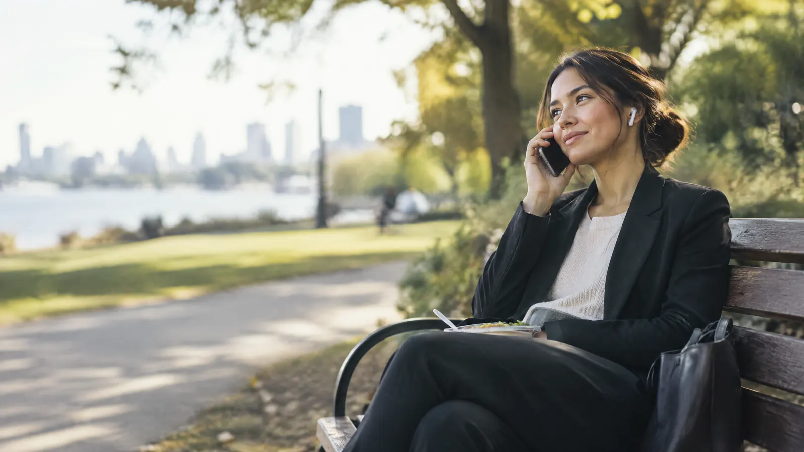 A young professional sitting on a park bench at lunch, calmly talking on the phone with earbuds, peaceful expression.