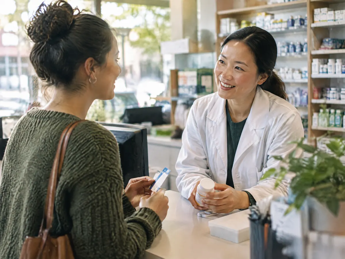 An employee at a neighbourhood pharmacy counter chats with a friendly pharmacist while picking up a prescription, a benefits card in hand.