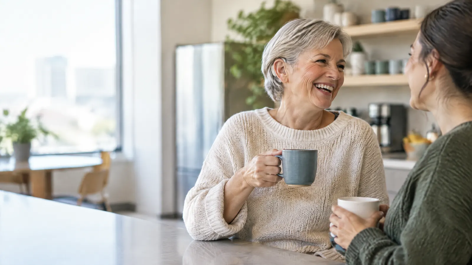 A smiling older woman chats with a younger coworker in a bright office kitchen, a small hearing aid visible behind her ear as she laughs over coffee.