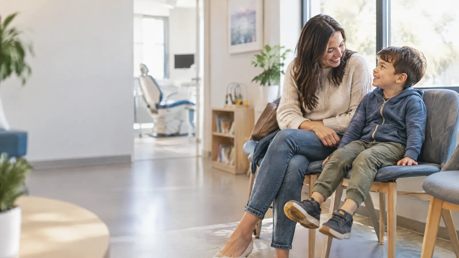 A mother and her young son sit side by side in a bright, modern dental clinic waiting area, smiling as he swings his feet beside her.
