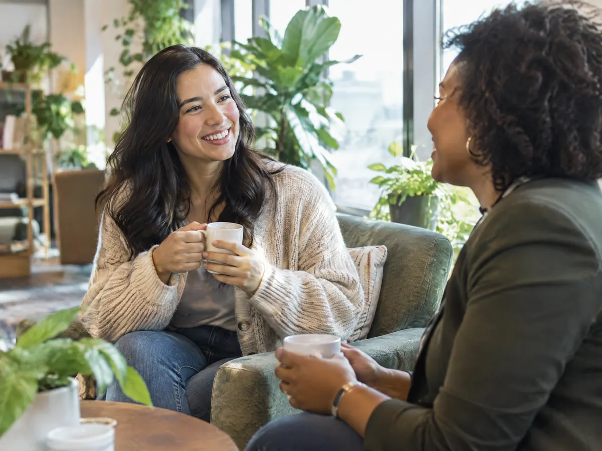 A woman in a cozy cardigan chats with a friendly HR colleague over coffee in a bright, plant-filled office lounge, both relaxed and smiling.