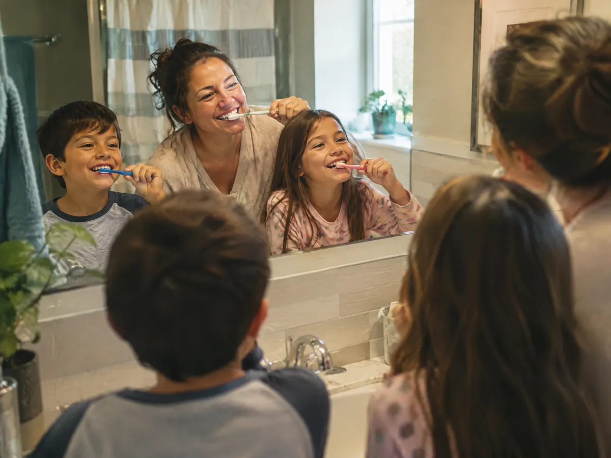 A parent and two school-aged kids brush their teeth together at a bathroom sink on a busy weekday morning, laughing in the mirror.