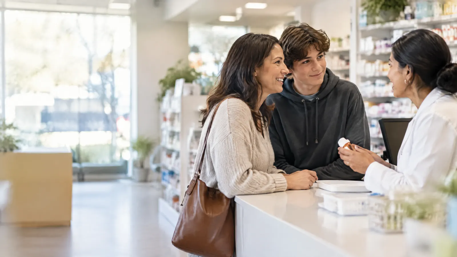 A mother and her teenage son chat with a friendly pharmacist at a bright neighbourhood pharmacy counter while picking up a prescription.