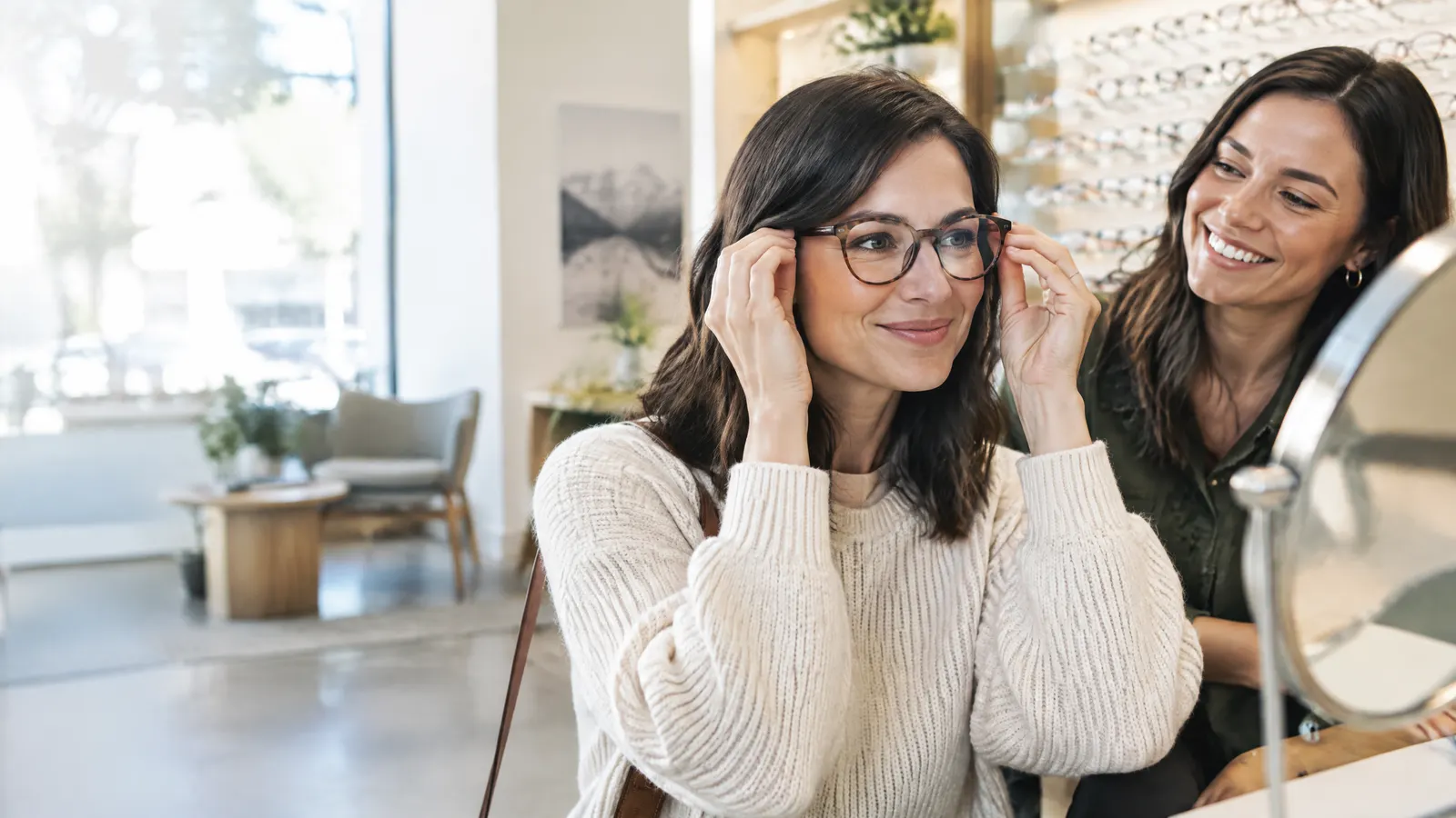 A woman in her thirties sits in a bright optometrist's shop trying on new eyeglass frames in a mirror while a friend smiles beside her.