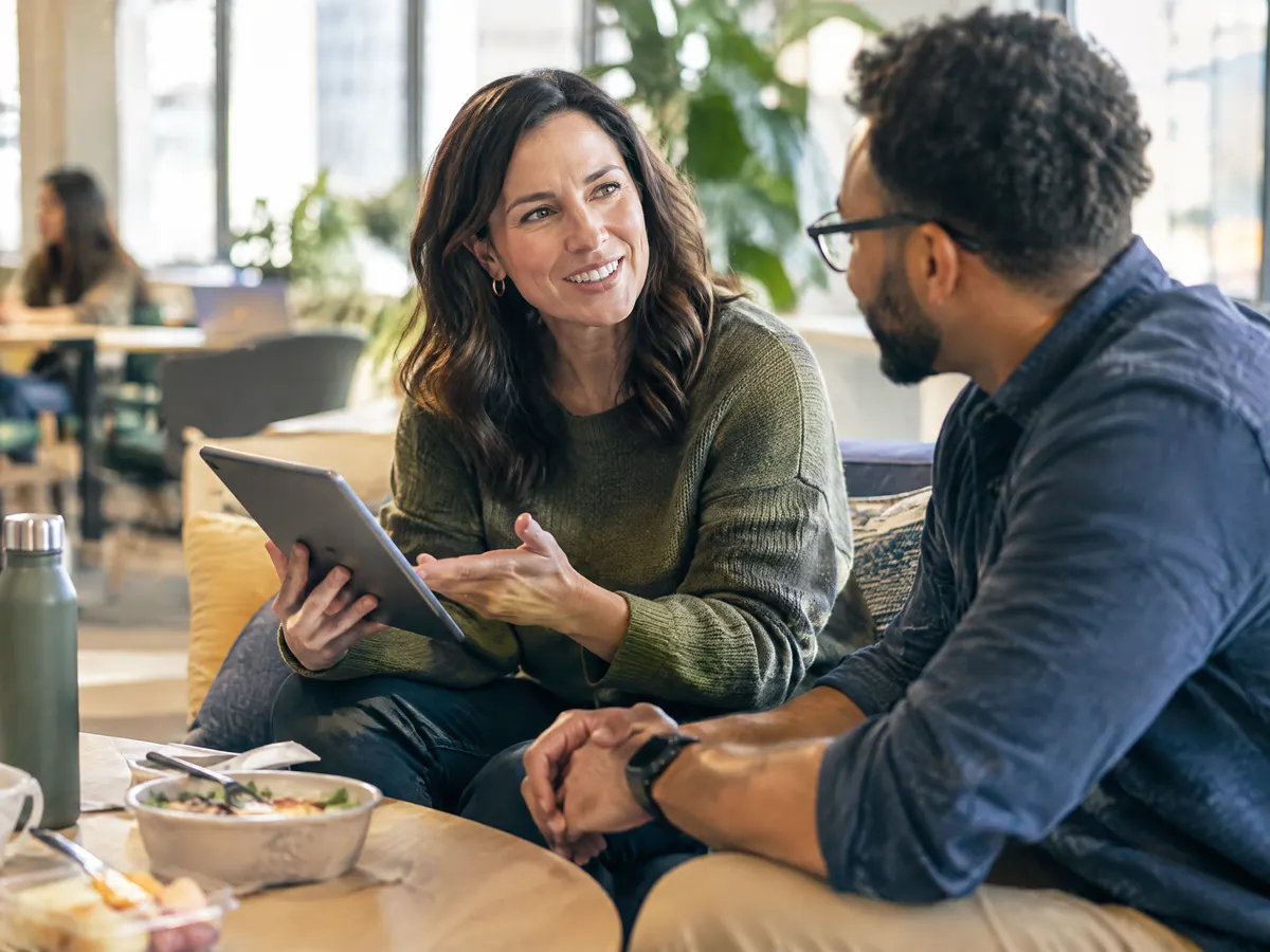 A mid-career employee chatting with a colleague in a bright, casual office lounge, holding a tablet and talking through workplace savings options over lunch.