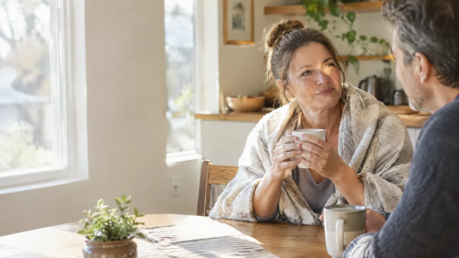 A woman in her forties sits at a sunny kitchen table with her partner, sharing coffee and a relaxed conversation during her recovery at home, a soft blanket over her shoulders.