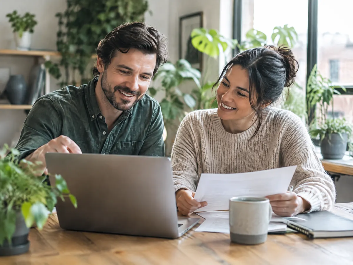 A small business owner and an employee chat warmly over a laptop in a bright, plant-filled office, reviewing paperwork together at a wooden table.