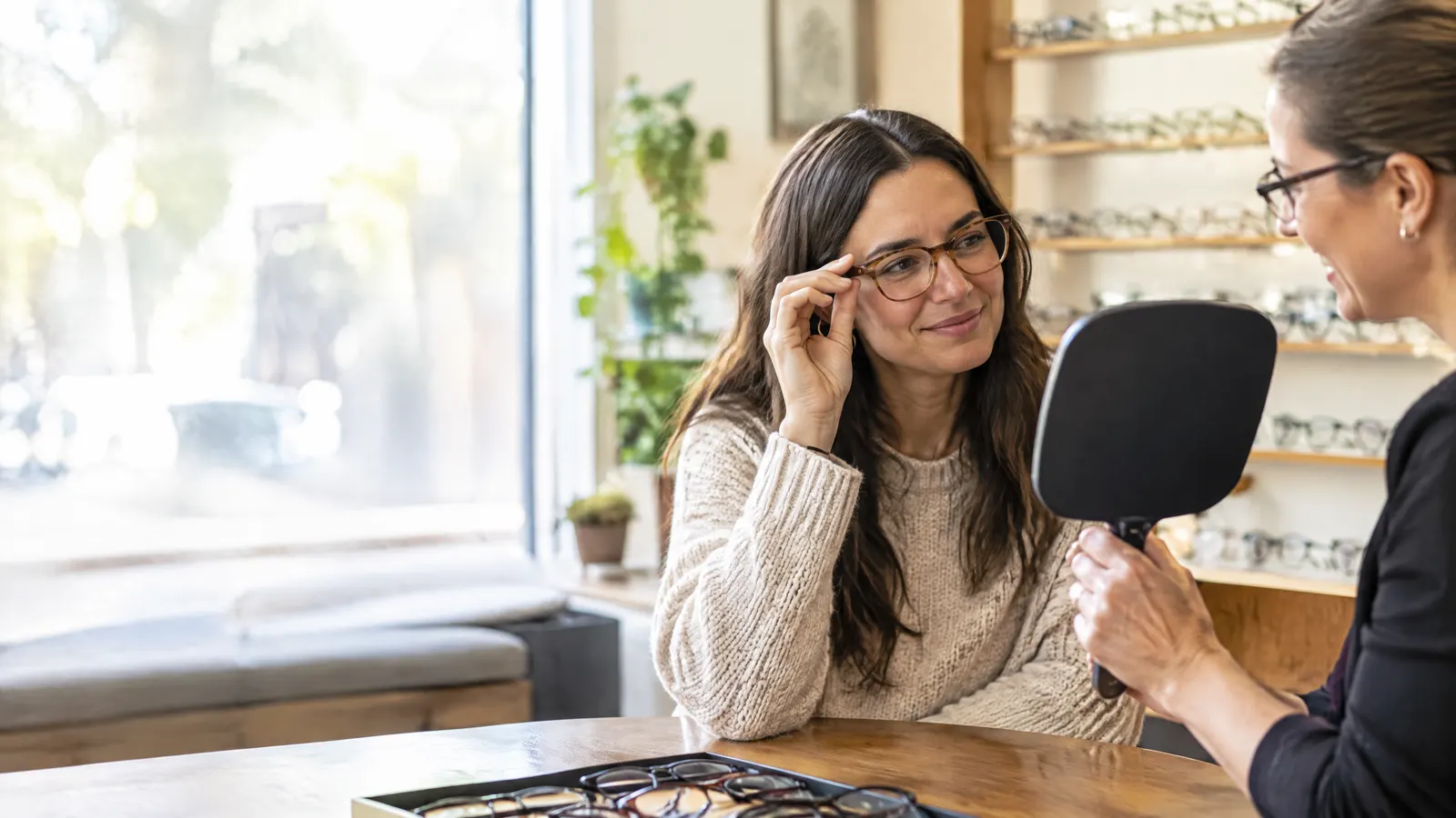 A woman in her thirties tries on new eyeglass frames at an optical shop counter while a friendly optician holds up a mirror, warm daylight through the window.