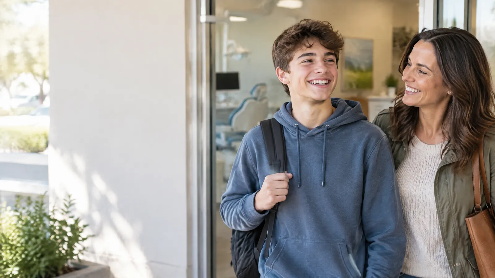 A mother and her teenage son leaving a bright dental office together, the son grinning to show off newly removed braces.