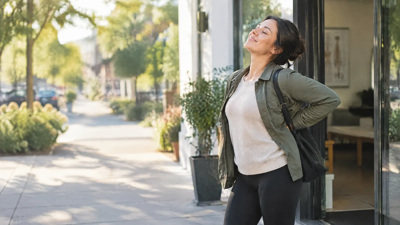 A woman leaving a chiropractor's clinic onto a sunny sidewalk, stretching her back happily.