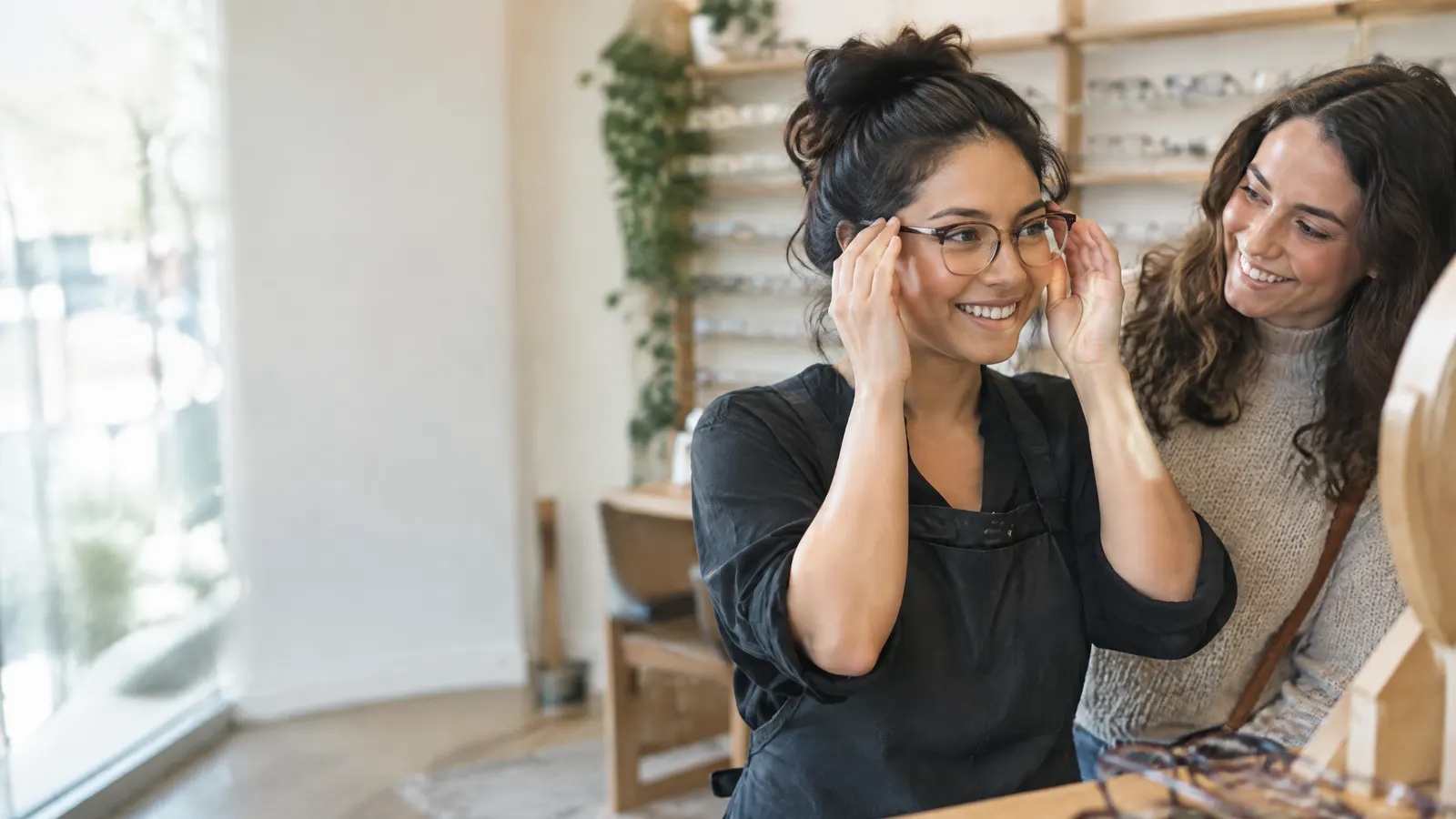 A retail worker trying on new eyeglasses at an optical shop with a friend.