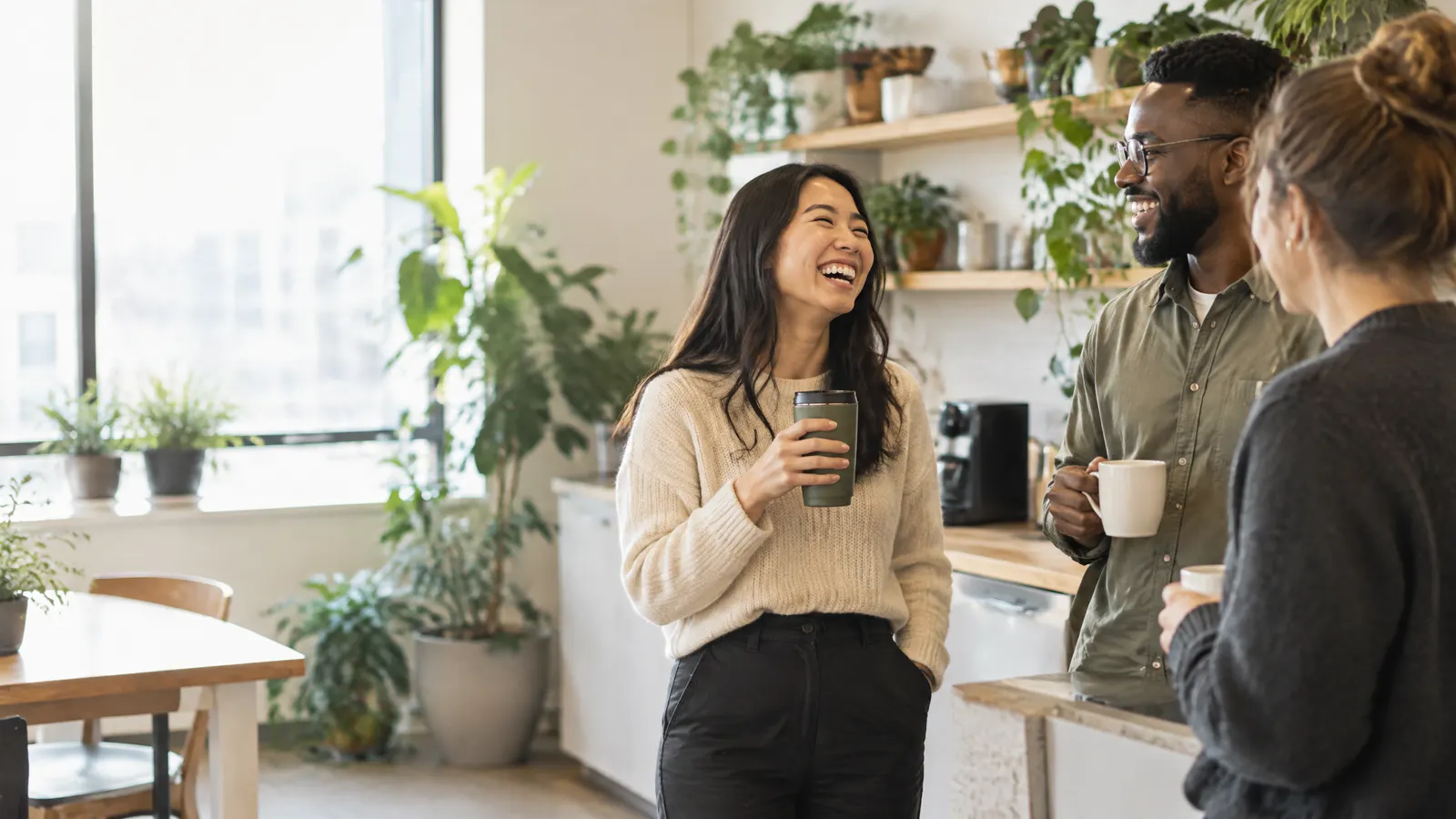 A small team of coworkers chatting over coffee in a bright, plant-filled office kitchen, one person laughing while holding a reusable mug.