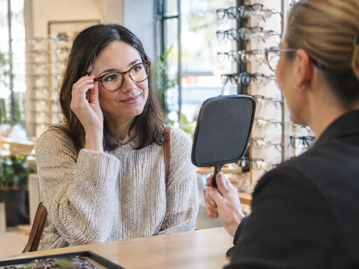 A woman in her thirties trying on new eyeglass frames at an optical shop counter while an optician holds up a small mirror.