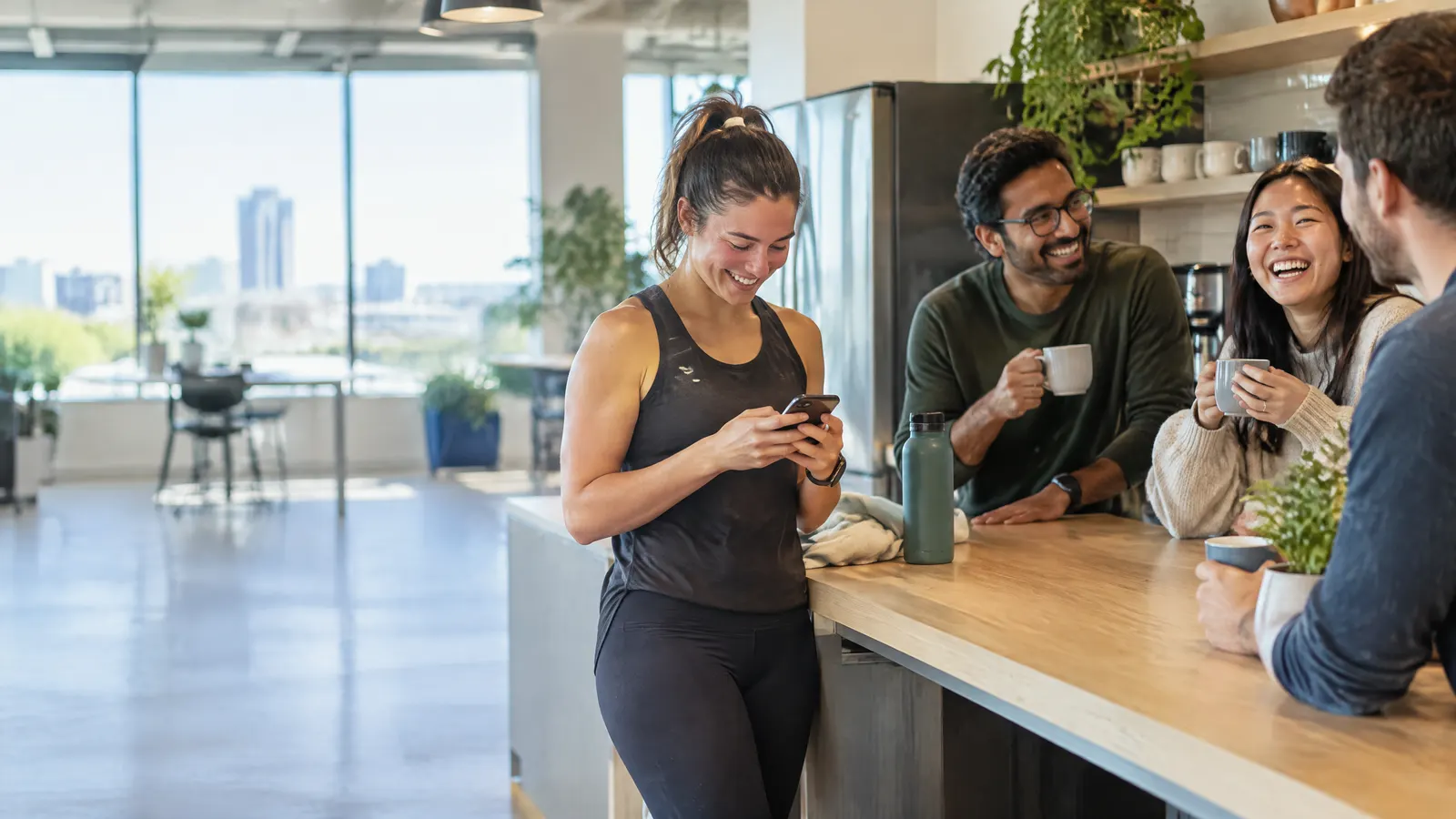 A small team at a bright Waterloo-style tech office laughs over coffee at a shared kitchen counter while one colleague checks her phone after a lunchtime run.