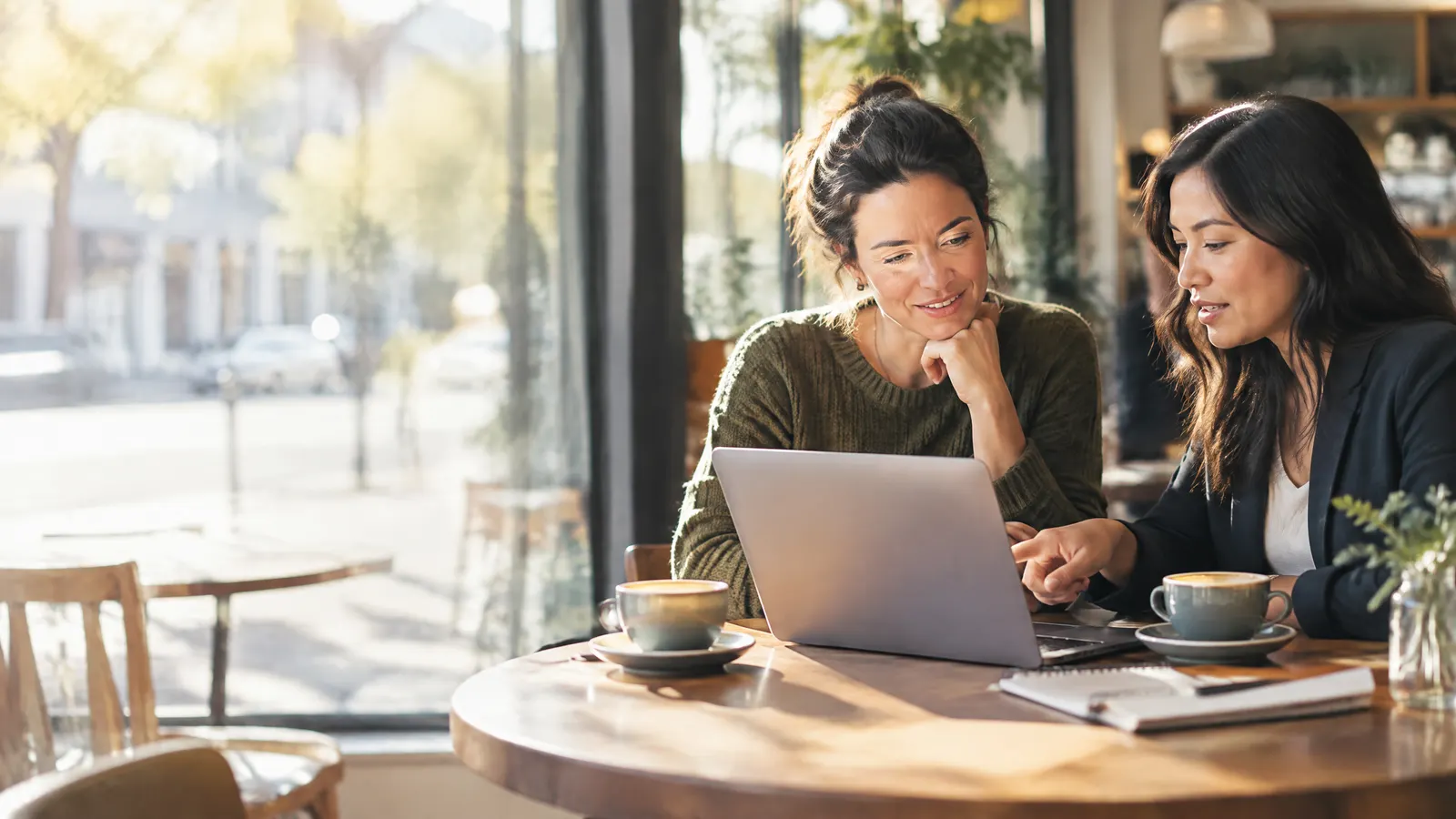 A small business owner and her HR lead sit at a sunny café table reviewing benefits options on a laptop, coffee cups and a notebook between them.