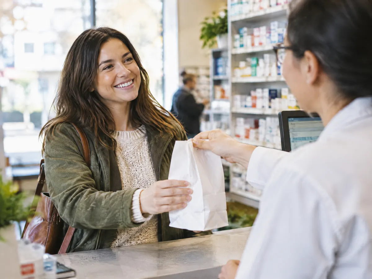 An employee smiles at a neighbourhood pharmacy counter as the pharmacist hands over a prescription bag.