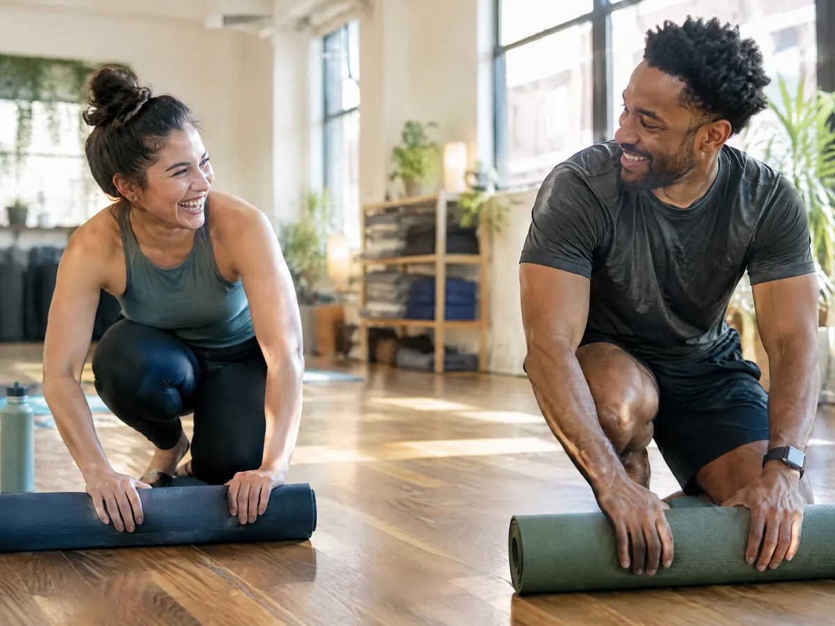 Two coworkers laugh while rolling out yoga mats in a bright community studio after work.