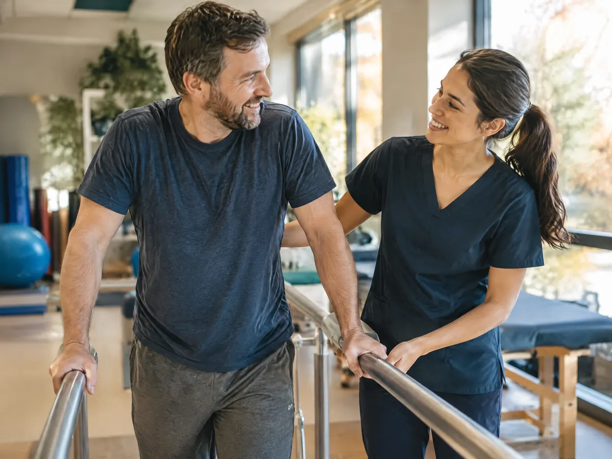 A man doing a light rehab session with a physiotherapist using parallel bars, both encouraging and smiling.