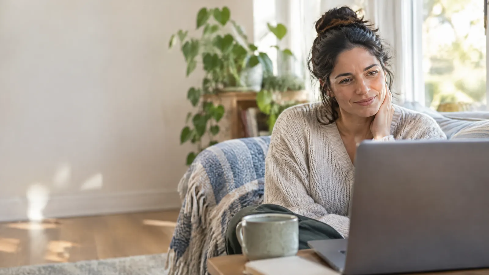 A woman in her thirties on a calm video session with a counsellor from her bright living room, a plant and a blanket nearby, at ease.