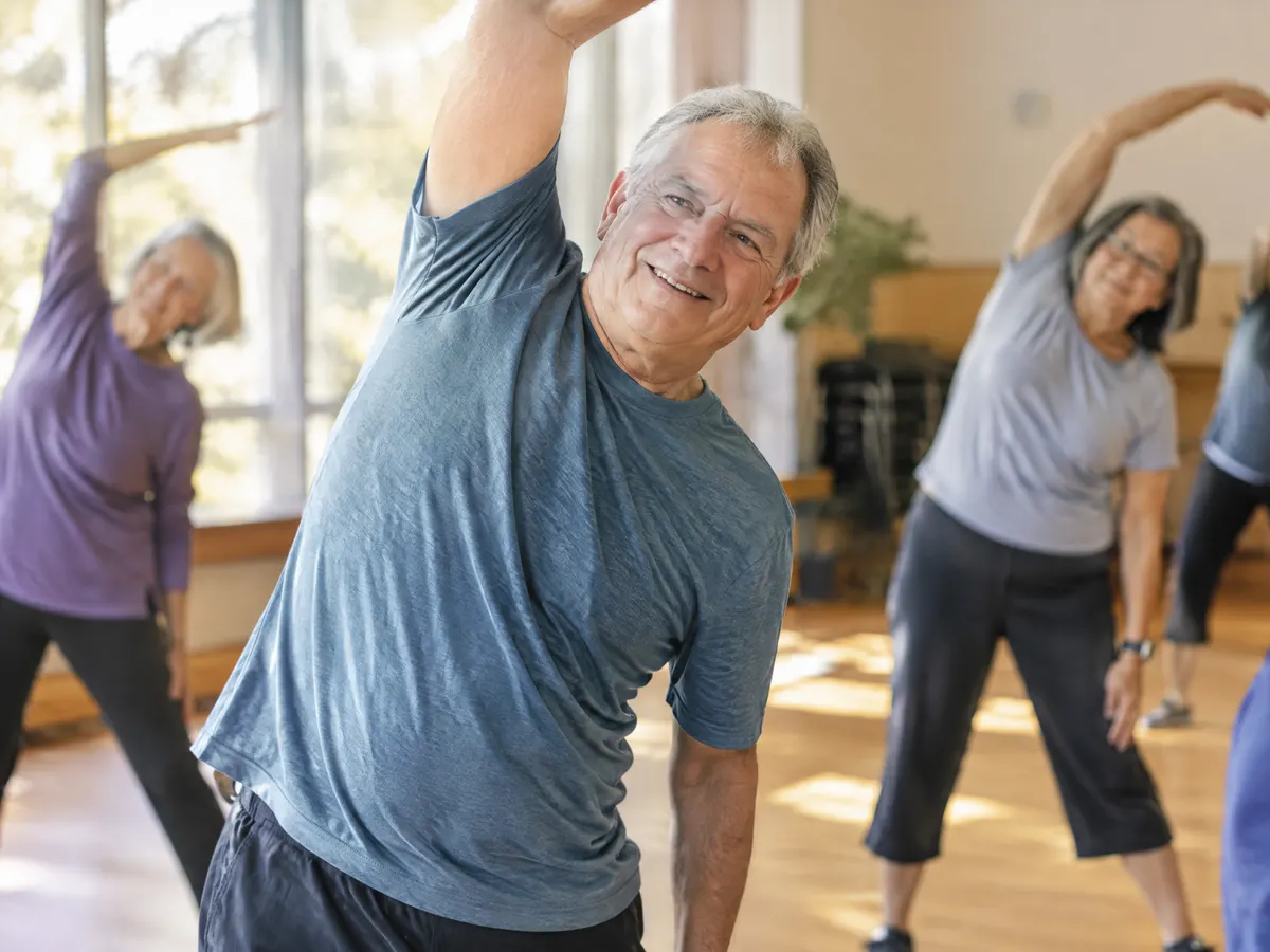 A man in his sixties doing a gentle stretching class at a community centre, smiling.
