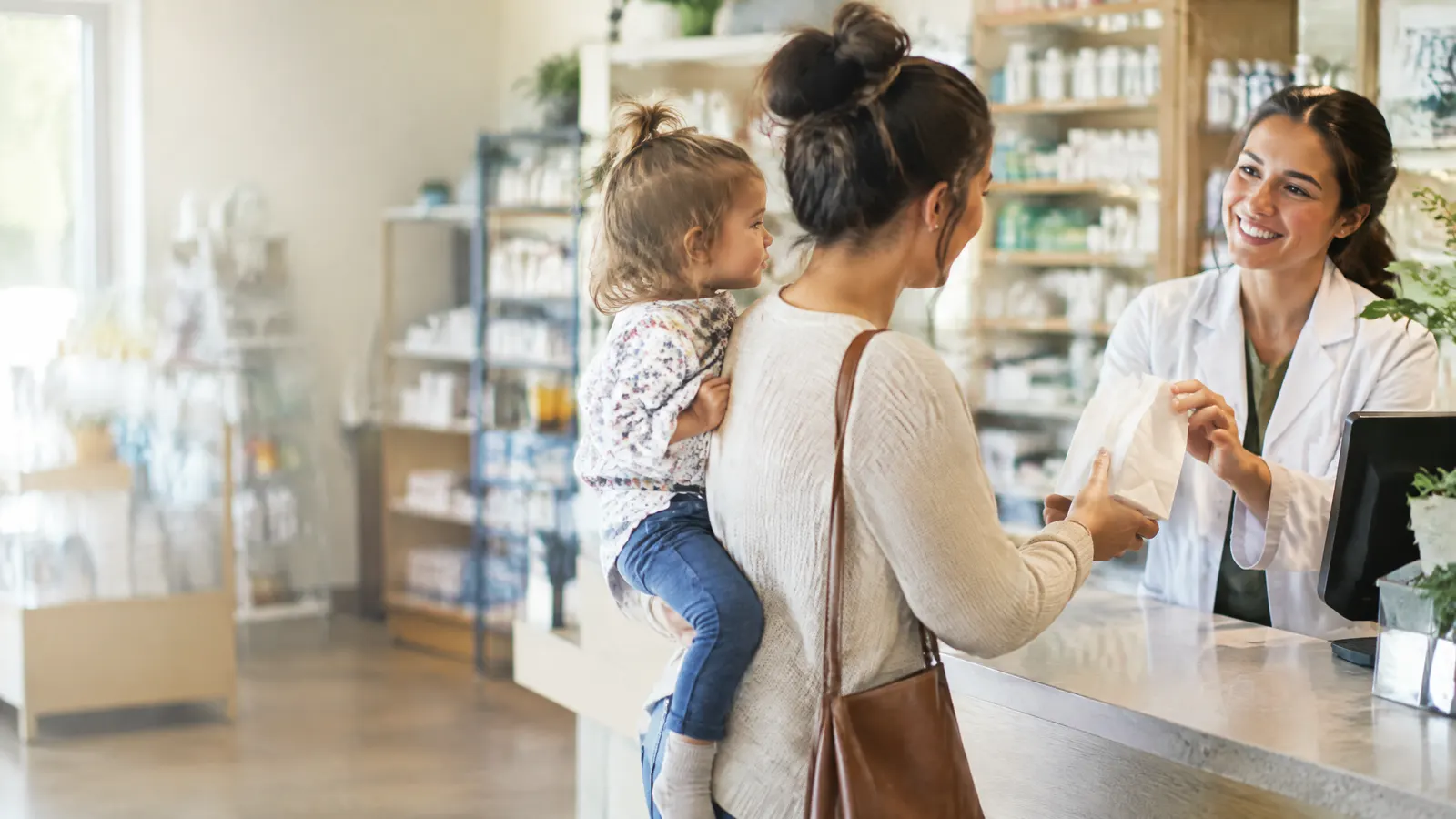 A young mother with a toddler on her hip picking up a prescription at a friendly neighbourhood pharmacy counter.