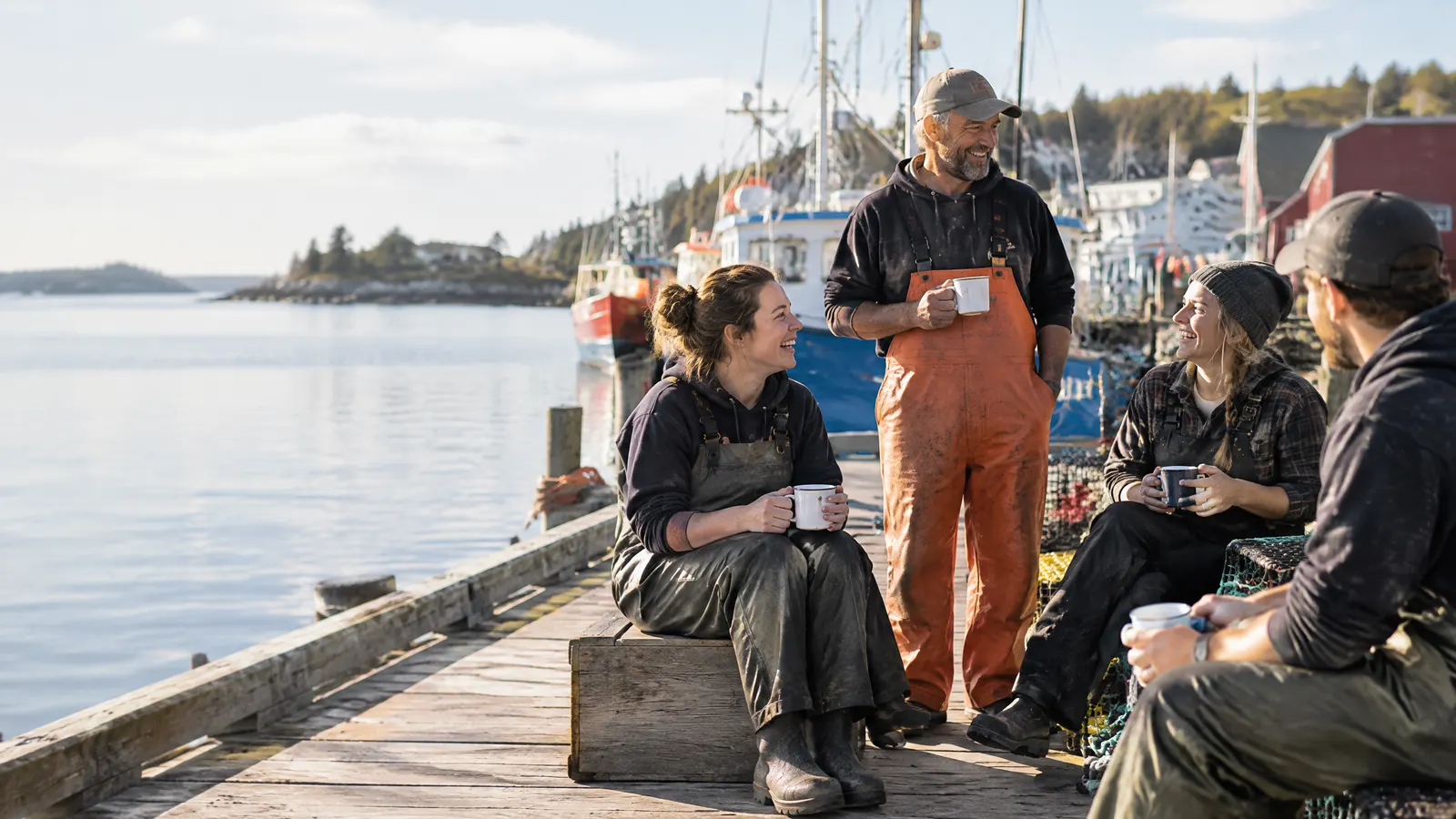 A small crew from a Nova Scotia family business shares coffee and laughs on a wooden wharf beside colourful fishing boats on a bright morning.