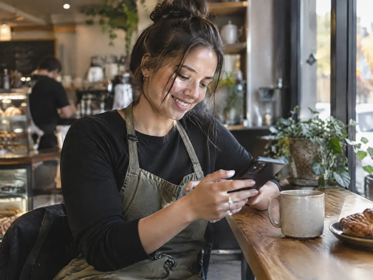 A young barista at a small cafe smiling as she books a dental cleaning on her phone during her break.