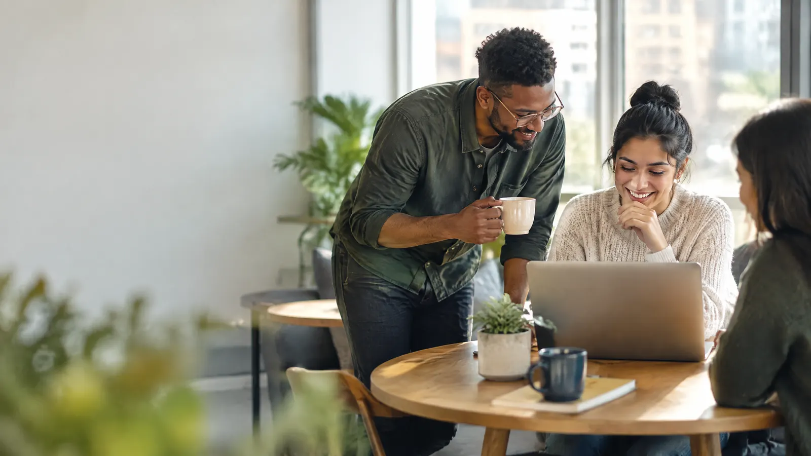 A small team gathers around a café-style table in a bright office, one person smiling while reading on a laptop as a coworker leans in with a coffee mug.