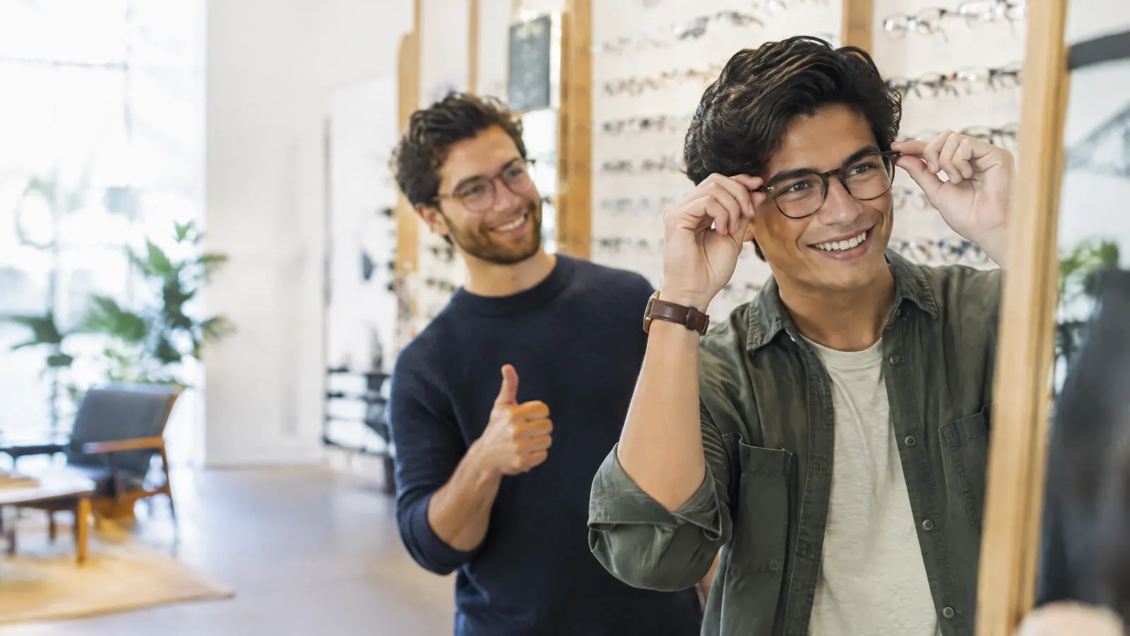A young man trying on new eyeglasses at a stylish optical shop, grinning at his reflection while a friend gives a thumbs up.